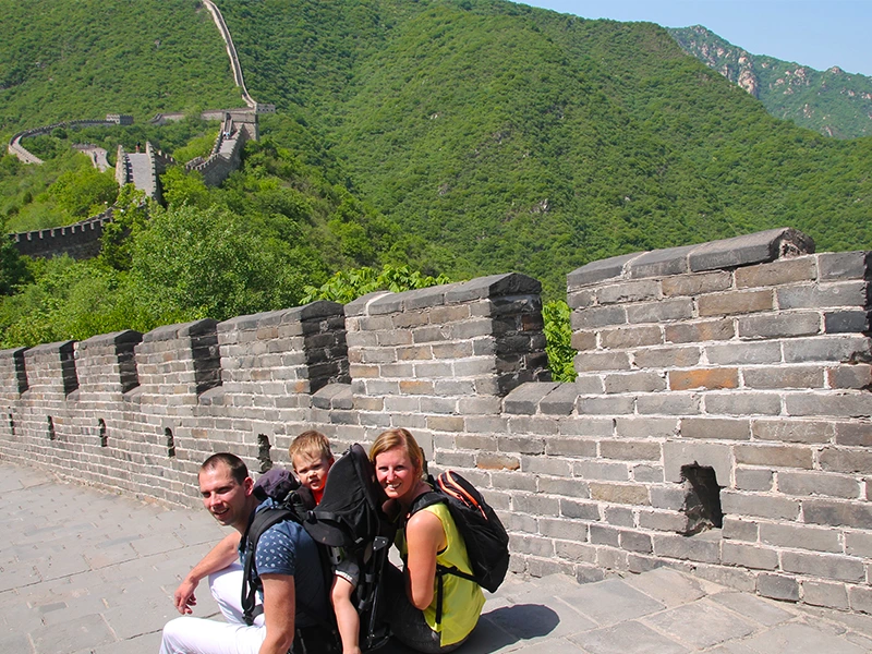 Gezin met een kind in een draagrugzak poseert op de Chinese Muur, met de groene heuvels en de slingerende muur op de achtergrond.