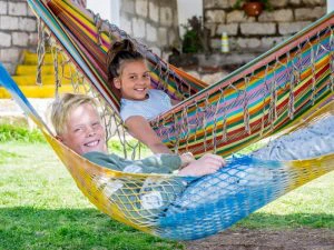 Twee kinderen liggen lachend in kleurrijke hangmatten in een tuin.