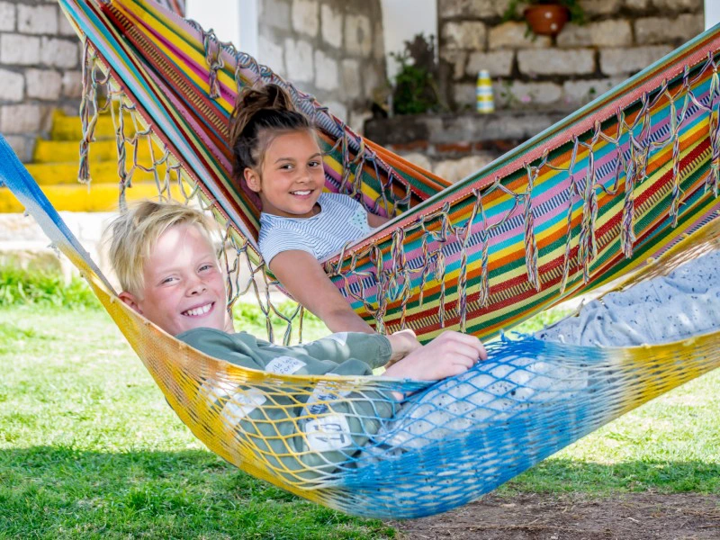 Twee kinderen liggen lachend in kleurrijke hangmatten in een tuin.