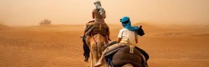 Een moeder op een kameel met een kindje daarachter die ook op een kameel zit. Ze rijden door een zandstorm in de Sahara van Marokko