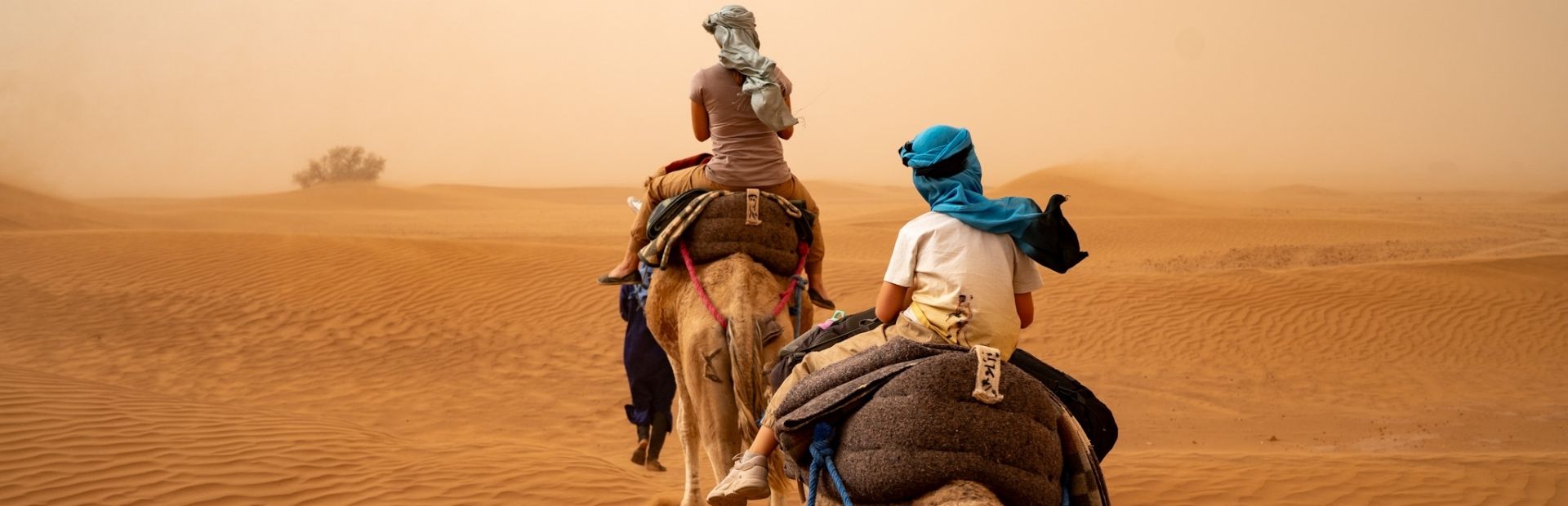 Een moeder op een kameel met een kindje daarachter die ook op een kameel zit. Ze rijden door een zandstorm in de Sahara van Marokko