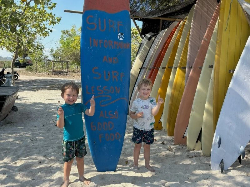 Twee jonge kinderen staan lachend op het strand in Kuta, Lombok, naast een groot blauw surfboard met opschrift “Surf information and surf lesson also good food”, met achter hen een rij gekleurde surfplanken.