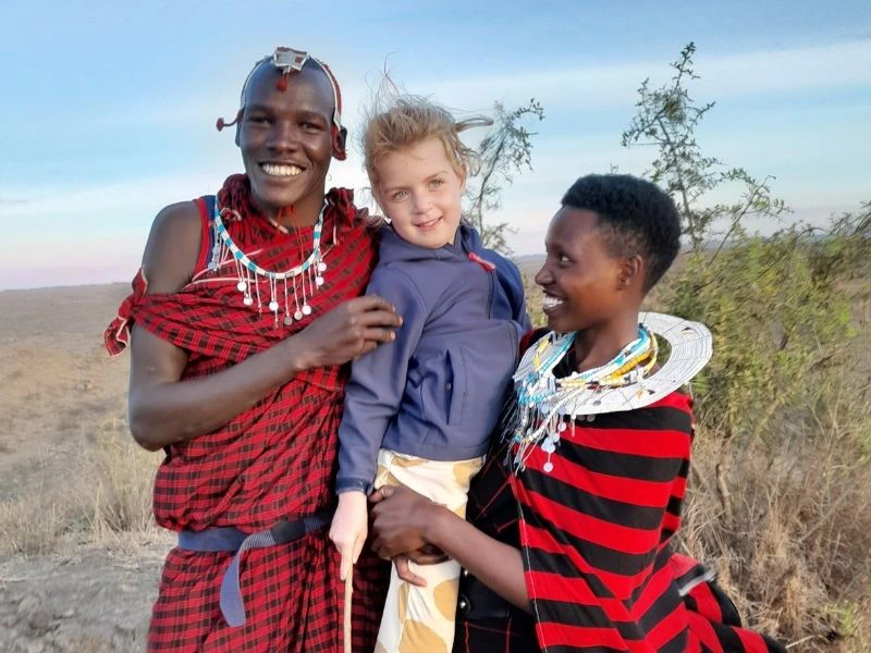 Twee Maasai in traditionele rood-zwarte kleding staan buiten in een savannegebied en houden een blond meisje lachend vast, met de blauwe lucht op de achtergrond.