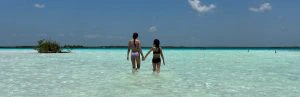 Twee kinderen lopen hand in hand door ondiep, helderblauw zeewater onder een zonnige lucht in Bacalar, Mexico.