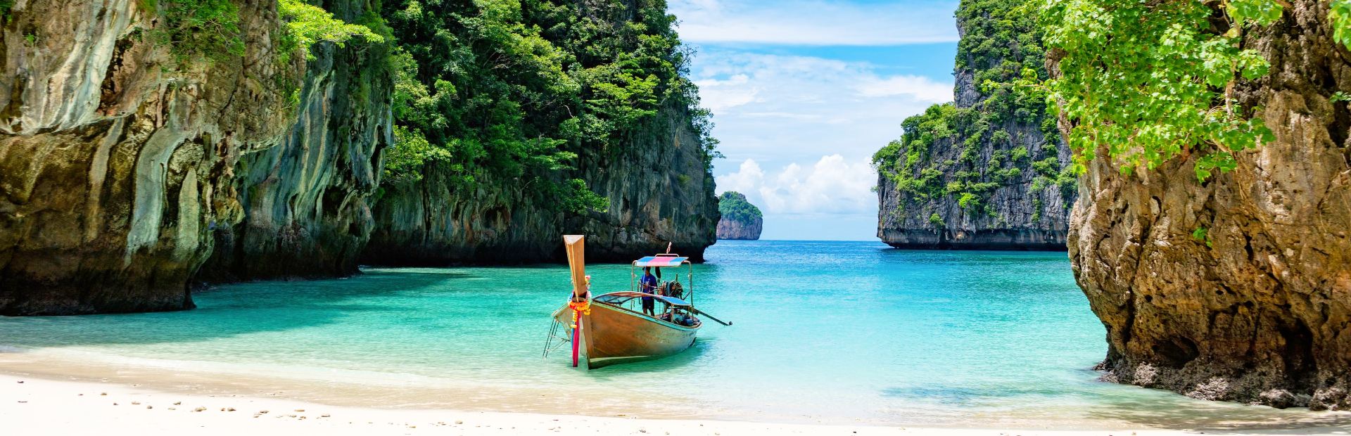 smalle boot van hout in een baai met rotsen rondom dobberend op een lichtblauwe zee met op de voorgrond een wit zandstrand