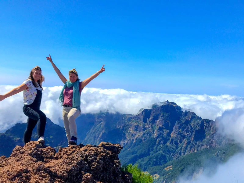 Twee vrouwen poseren vrolijk op een bergtop met uitzicht op ruige bergen en een wolkendek.
