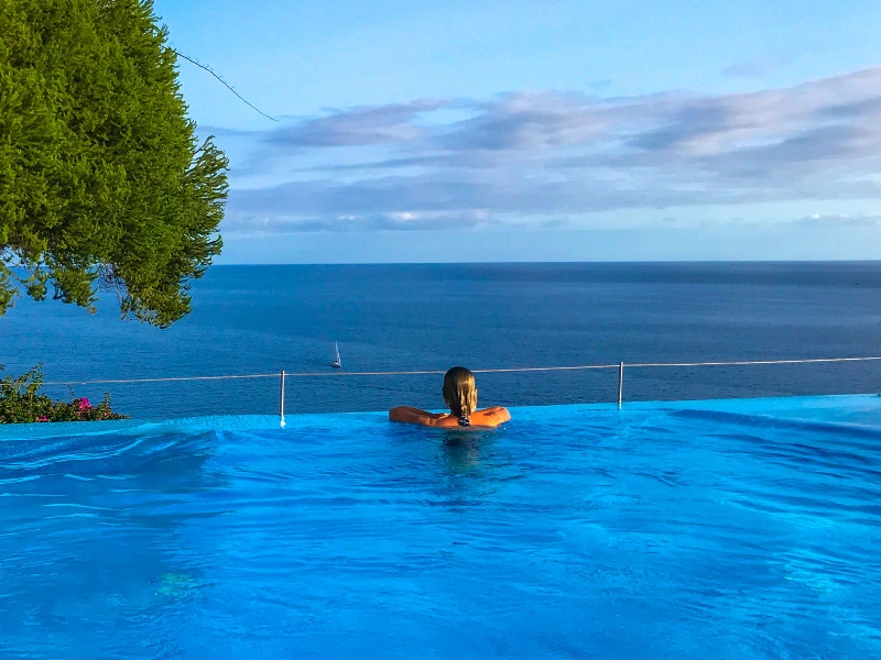 Vrouw ontspant in een infinity pool met uitzicht op de zee en een zeilboot aan de horizon.