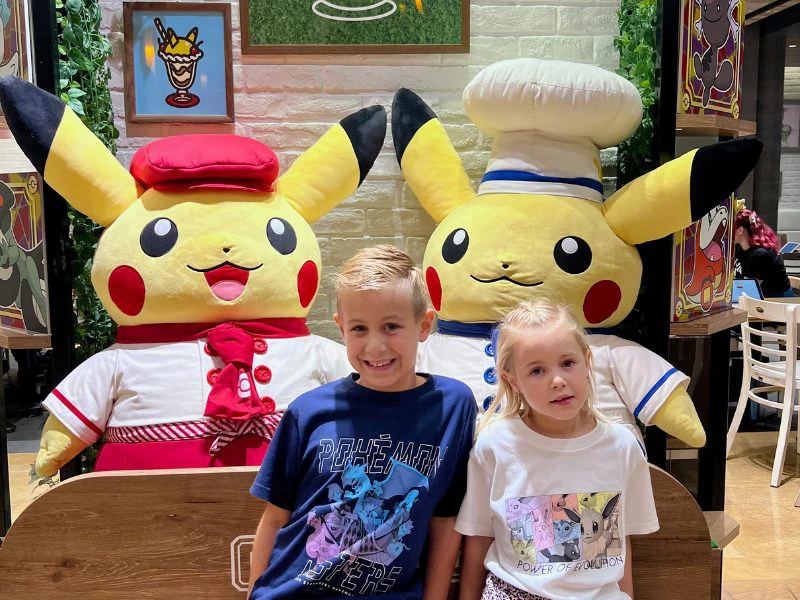 Een jongen en meisje met anime shirts aan zitten op een houten bankje in een Pokemoncafe in Tokyo met achter hun twee grote, gele Pikachu's: links als serveerder in het rood en rechts als kok in het blauw en wit met koksmuts.