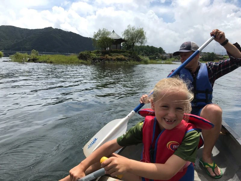 Vader met dochter zitten in een kano op een meer in Japan. Het meisje heeft een rood zwemvest aan en de man een blauwe. Achter hen is een bergachtig en groen landschap en de lucht kleurt blauw met voornamelijk witte wolken overal.