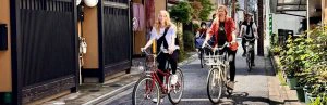 Moeder en twee tieners fietsen door een smalle straat in Kyoto, Japan. Ze rijden langs traditionele houten huizen en groene planten. De zon schijnt en de sfeer is ontspannen en vrolijk.
