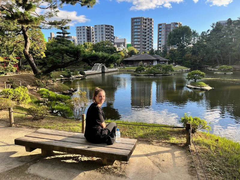 Reizigster met bruin haar zit op een houten bankje bij een stadspark met water en veel groen en in de verte hoge gebouwen en een blauwe lucht met witte wolken hier en daar.