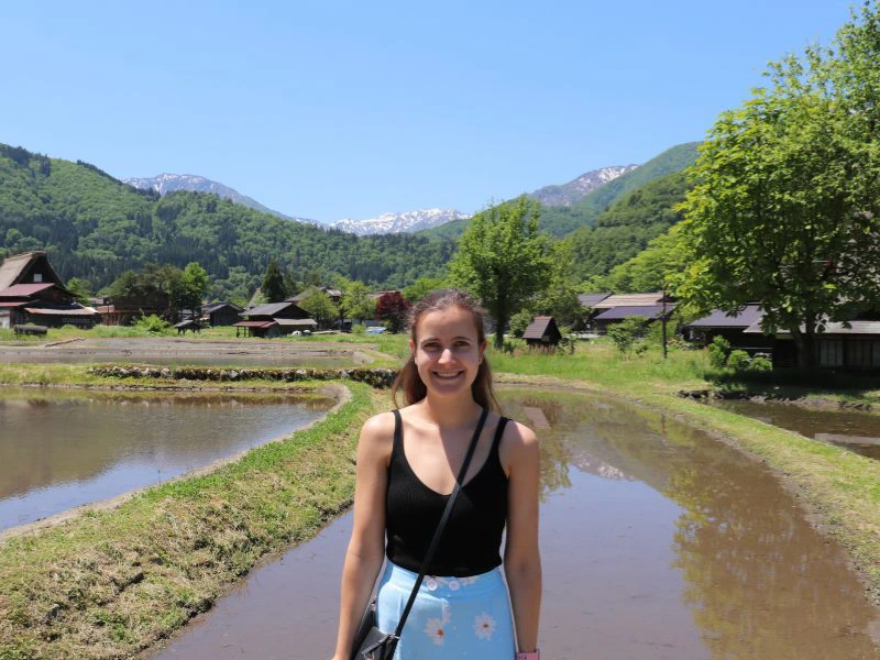 Reizigster met lang bruin haar staat lachend bij een natte rijstterrassenveld in een bergdorp in Japan. Ze draagt een zwarte top en lichtblauwe rok met bloemenprint. Op de achtergrond traditionele Japanse houten huizen met schuine daken, groene heuvels en besneeuwde bergtoppen onder een heldere blauwe lucht.