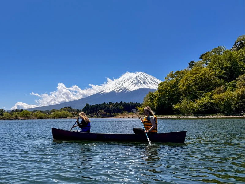 Moeder en dochter peddelen in een open kano op een blauw meer, met op de achtergrond de besneeuwde witte top van Mount Fuji onder een helderblauwe lucht met witte wolken. De moeder draagt een felgeel reddingsvest en een grijsgroen T-shirt, de dochter een blauw reddingsvest en zwarte kleding. Rond het meer staan groene bomen en lage bebouwing, met lichte heuvels op de achtergrond.