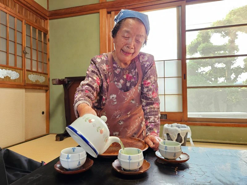 Japanse oude vrouw met bloemetjes jurk en bloemetjesschort die thee inschenkt in drie witte kopjes die op houten onderzetters staan. De theepot is ook wit met bloemen.