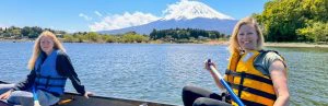 oeder en dochter zitten in een open kano op een blauw meer, met op de achtergrond de besneeuwde witte top van Mount Fuji onder een helderblauwe lucht met witte wolken. De moeder draagt een felgeel reddingsvest en een grijsgroen T-shirt, de dochter een blauw reddingsvest en zwarte kleding. Rond het meer staan groene bomen en lage bebouwing, met lichte heuvels op de achtergrond.