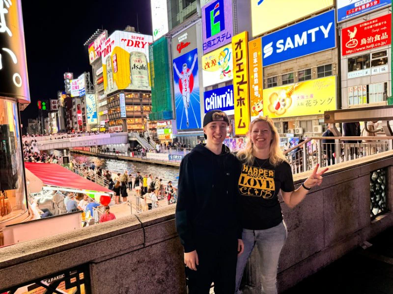 Moeder met zoon links van haar met pet op en zelf met een peaceteken met haar vingers staan voor een stenen muurtje met achter hen de drukke straat van Osaka met veel neon reclameborden.