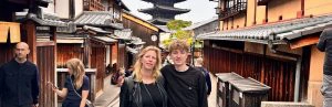 Moeder met haar zoon staan tegen elkaar aan te poseren in een straat van Kyoto. Om hen heen staan ook andere mensen. De huizen zijn van hout, geel en met dakpannen bedekt. Aan het einde van de straat torent een tempel er bovenuit.