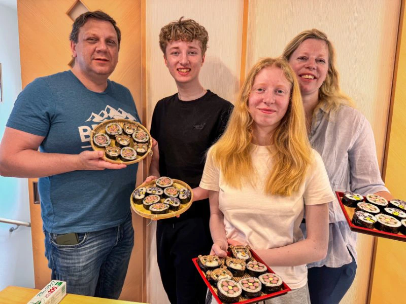 Vier personen, twee volwassenen en twee tieners, staan binnen en houden borden met zelfgemaakte sushi vast. In de sushi zijn figuren te zien zoals bloemen en beertjes. Ze staan voor een wand met lichte houten panelen.