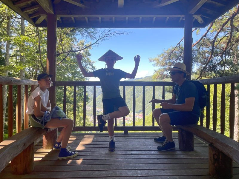 Vader en dochter zitten in een Japans uitkijkpaviljoen terwijl de zoon in het midden op één been poseert met een rieten hoed. Op de achtergrond zijn bergen, bos en een felblauwe lucht te zien.