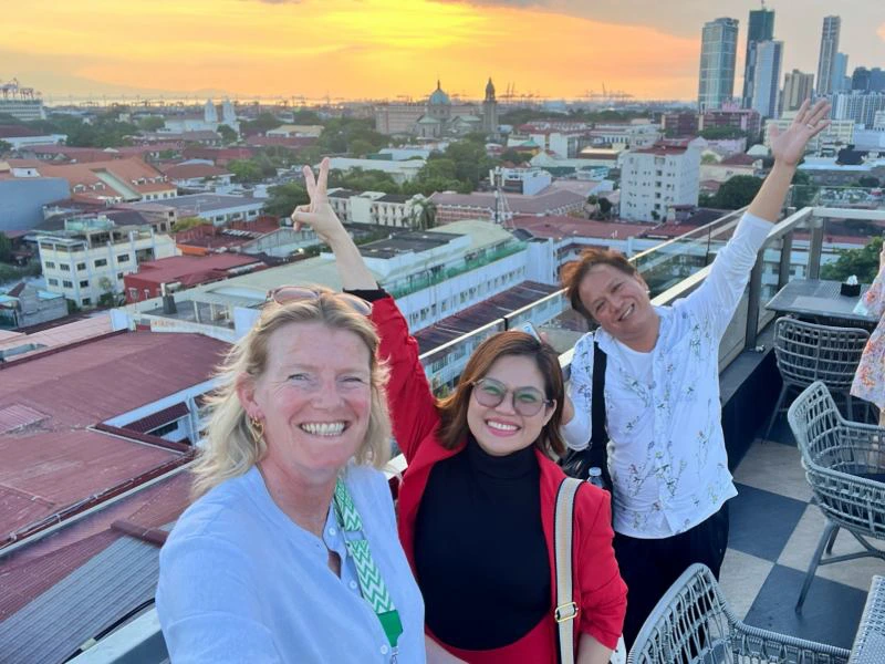 Drie lachende mensen poseren op een dakterras met uitzicht over Manila bij zonsondergang