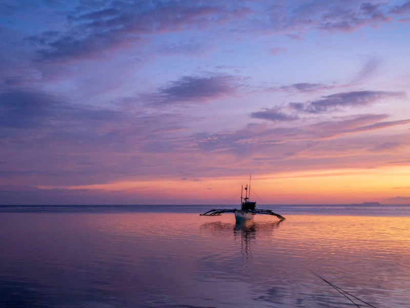 de lucht is gekleurd van paars naar geel en een bootje dobbert op de branding tijdens de zonsondergang op Siquijor