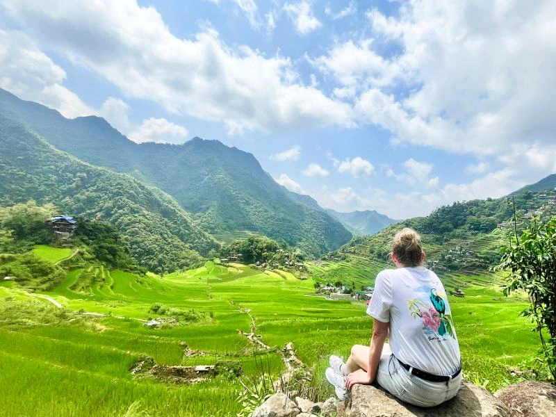 Persoon in wit T-shirt zit op een steen en kijkt uit over groene rijstterrassen en bergen onder een deels bewolkte hemel