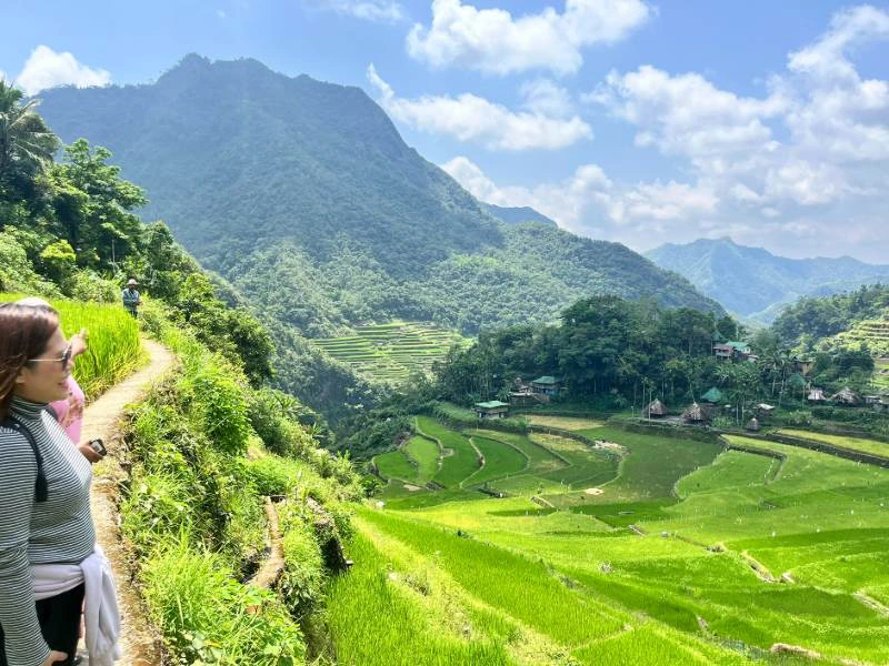 Wandelaars op een smal pad langs felgroene rijstterrassen, met uitzicht op een vallei en beboste bergen onder een blauwe lucht met wolken