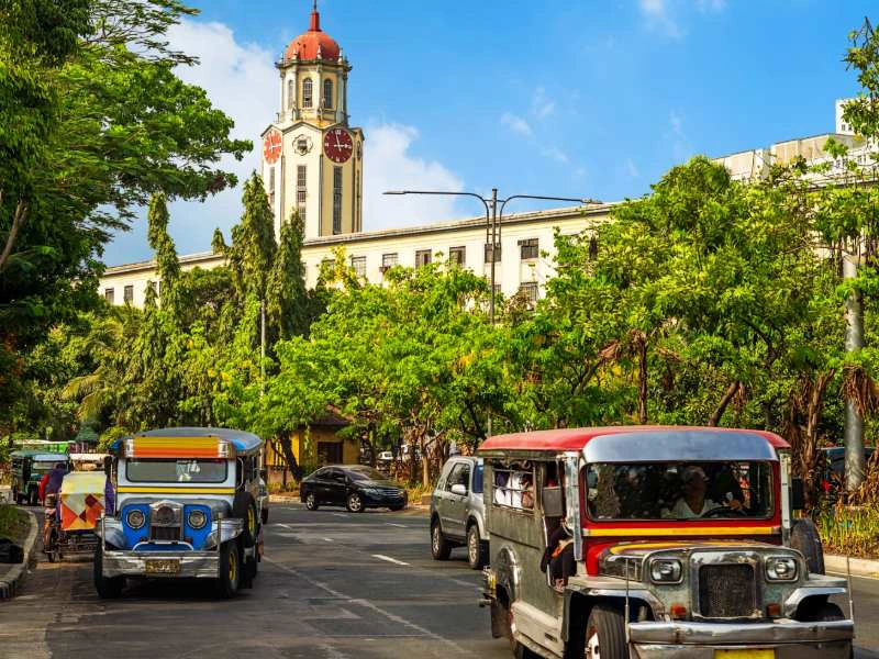 Straatbeeld met traditionele Filipijnse jeepneys voor een historisch gebouw met klokkentoren, omgeven door groene bomen en blauwe lucht.