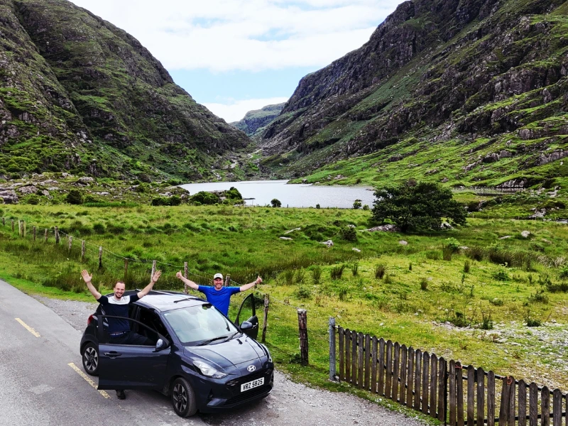 Twee mannen staan bij hun auto met armen omhoog in een nationaal park in Ierland